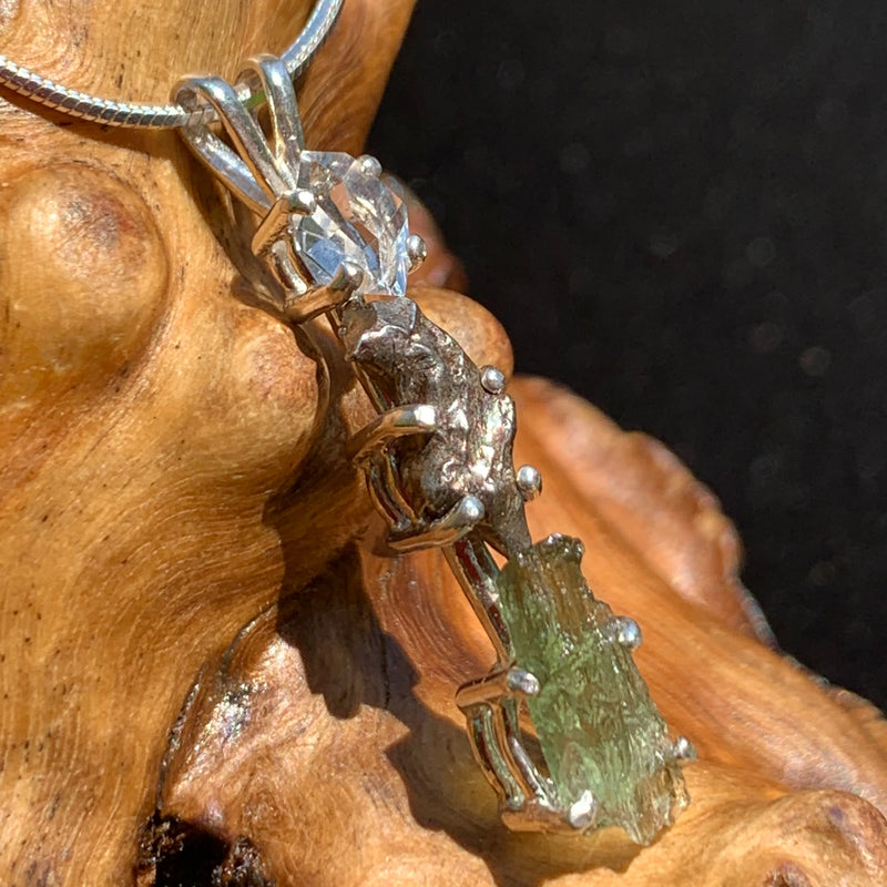 side view of a sterling silver pendant necklace with Herkimer diamond, moldavite, and campo del cielo meteorite sitting on driftwood for display