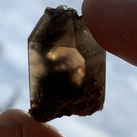tiny brookite crystals on a smokey quartz point held in hand up to the sky with sunlight shining through
