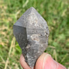medium and tiny brookite crystals on a smokey quartz point held in hand with sun shining on it against a grassy background