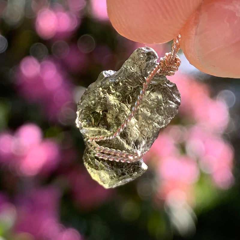 Moldavite sterling silver pendant  in the sun with blurry pink flowers in the background