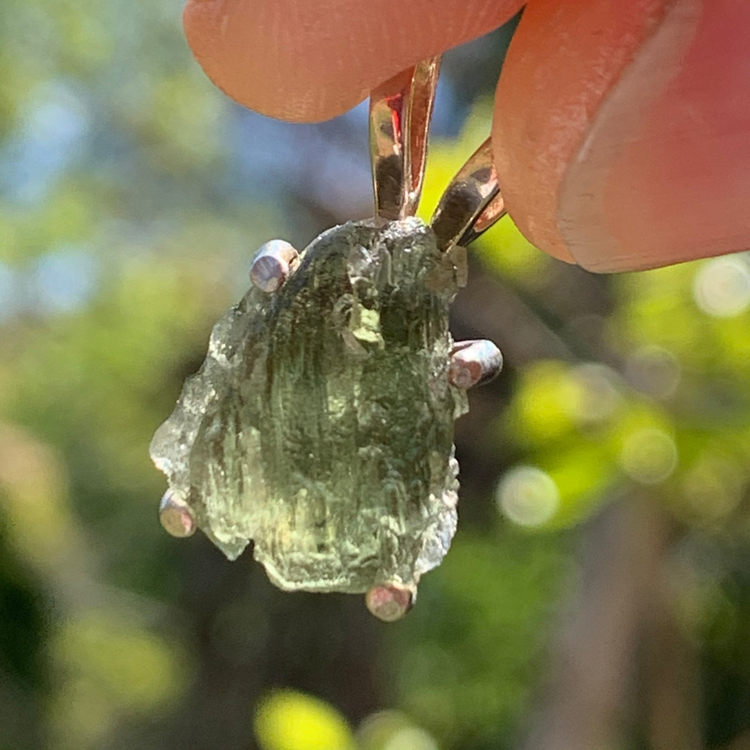 raw moldavite tektite in 4 prong sterling silver basket pendant held in hand with sunlight shining through it