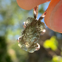 raw moldavite tektite in 4 prong sterling silver basket pendant held in hand up to sky with sunlight shining through