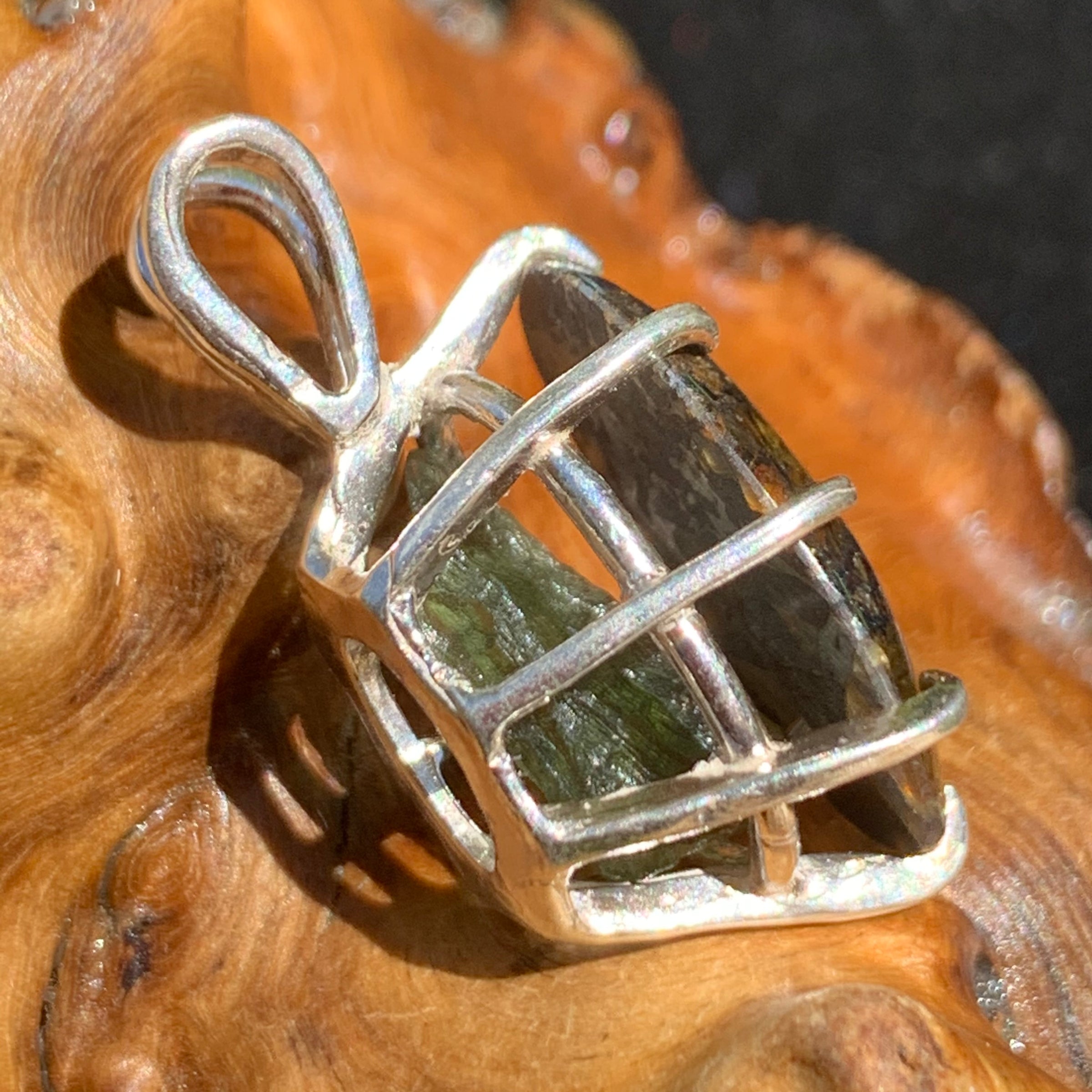 moldavite shows through the side of a sterling silver sericho pallasite meteorite and raw moldavite tektite basket pendant sitting on driftwood for display
