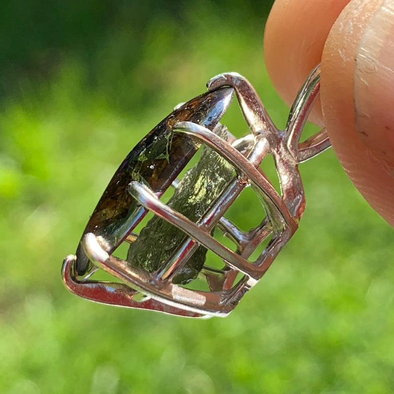 moldavite shows through the side of a sterling silver sericho pallasite meteorite and raw moldavite tektite basket pendant held up on display to show details