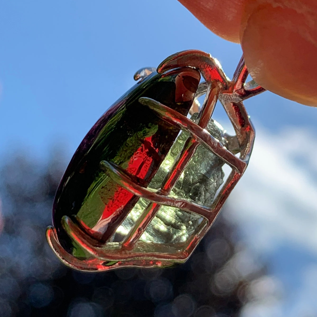 sterling silver sericho pallasite meteorite and raw moldavite tektite basket pendant held up on display with sunlight shining through to show details