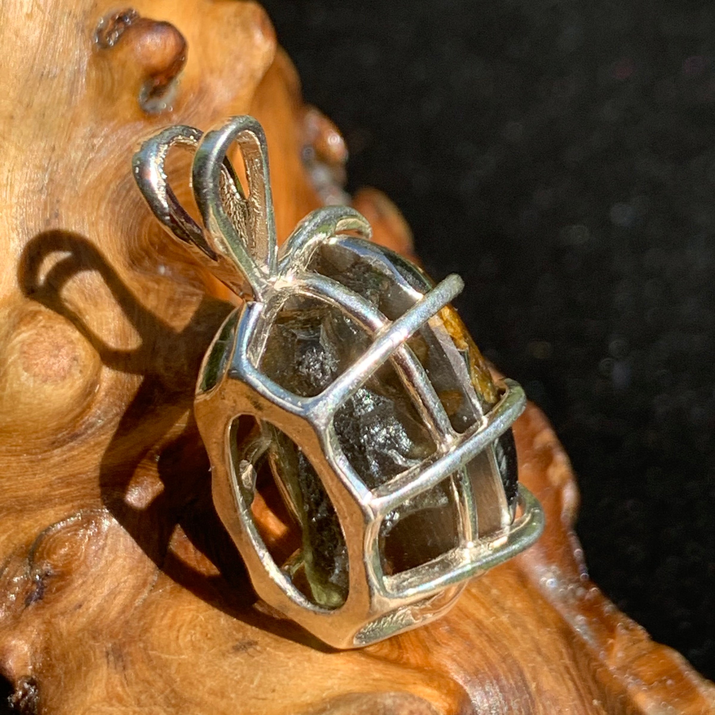 moldavite showing through the backside of a sterling silver sericho pallasite meteorite and raw moldavite tektite basket pendant sitting on driftwood for display