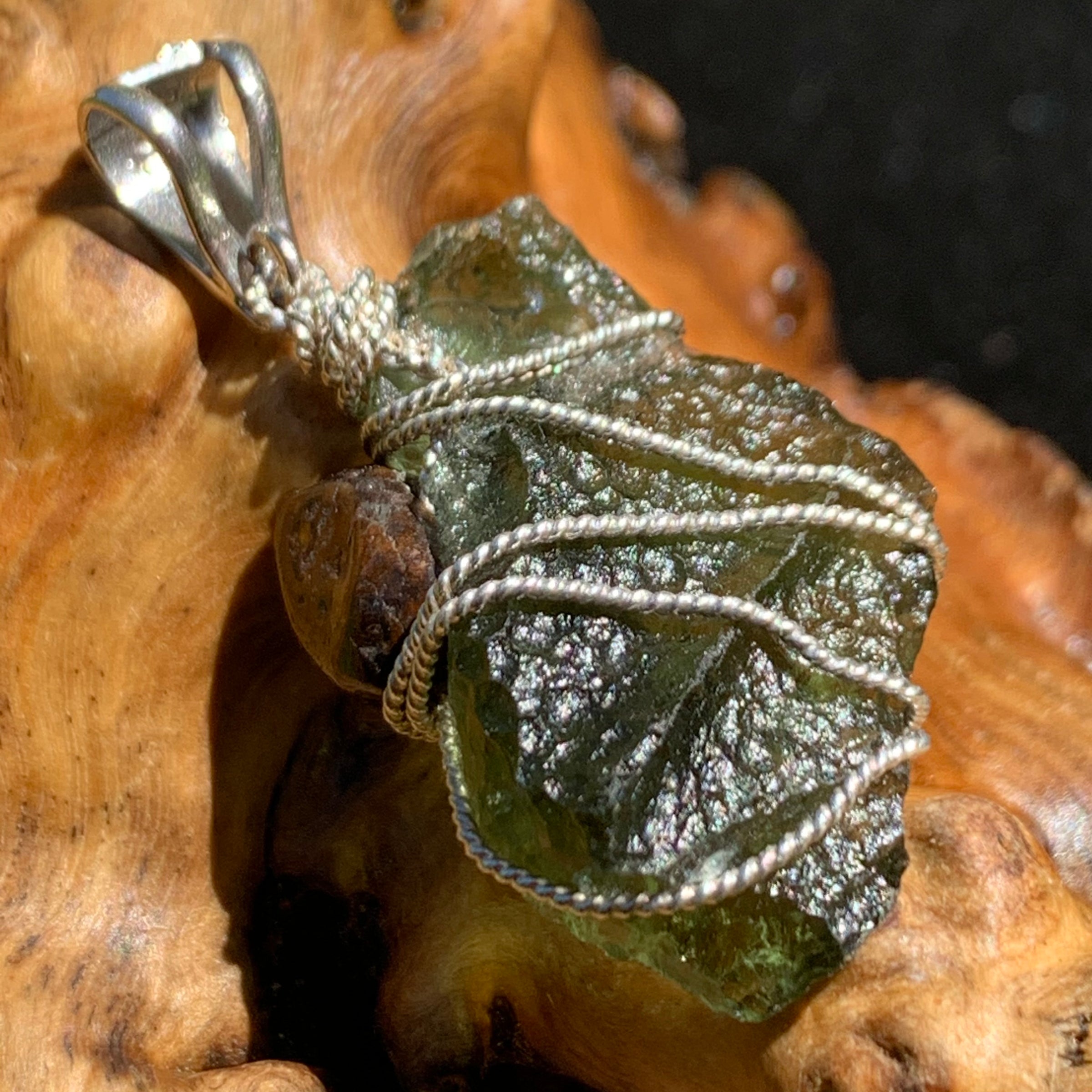 backside close up of a raw moldavite tektite and nwa 869 meteorite bead sterling silver wire wrapped pendant sitting on driftwood for display