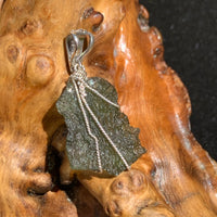 backside of a raw moldavite tektite and black tourmaline sterling silver wire wrapped pendant sitting on driftwood for display